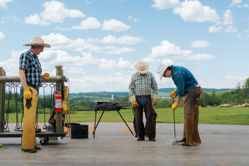 Three men are on a wooden deck outside, under a blue sky with puffy clouds and mountains in the background. One of them is branding a piece of wood, and they've got more irons in fire. 