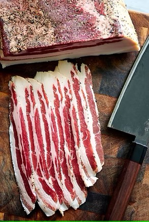 Slices of homemade Berskshire Pork Cured Bacon from above, laid out on a wooden cutting board. The chunk they were cut off of and a cleaver are above and beside the slices.