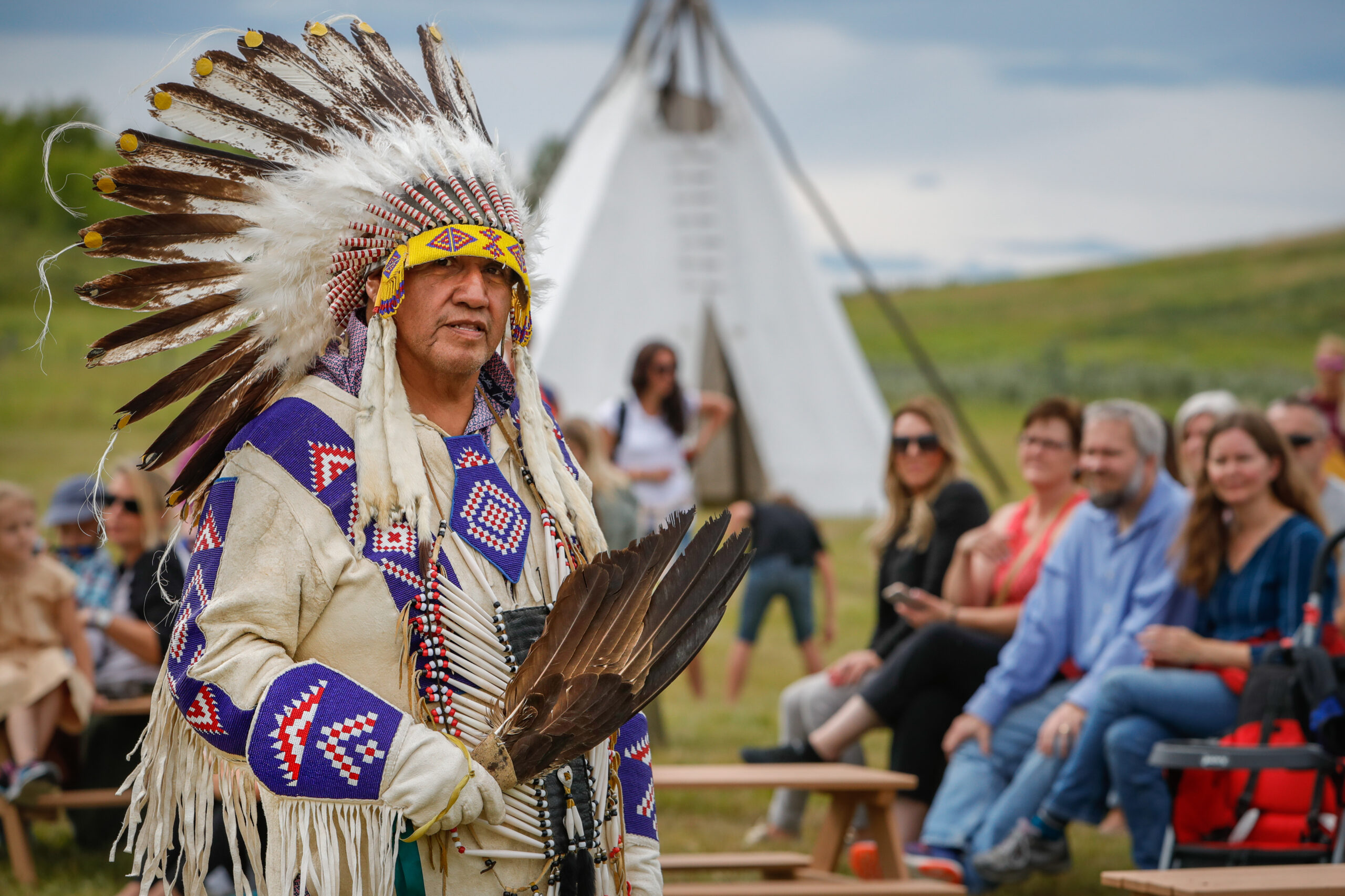 Indigenous Days. An indigenous man in regalia stands in front of a teepee with people around, and green grassy hills in the background.