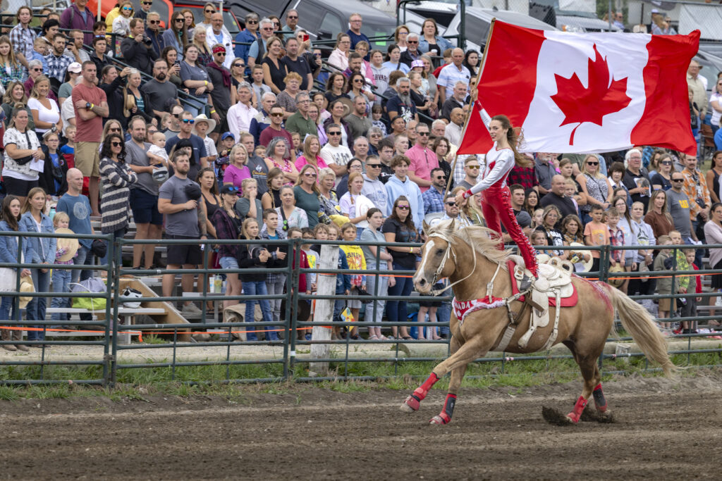 Cochrane Lions Rodeo - a crowd watches from the bleachers. A woman is riding a horse, standing up, holding a Canadian flag. Her and the horse are dressed in red and white.