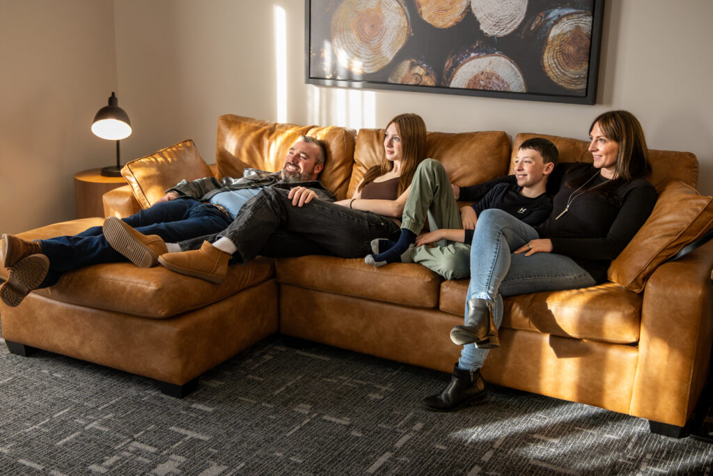 A family reclines on a brown leather sofa in dappled sunlight. There is a large piece of art on the wall above them - a photograph of the ends of logs showing a variety of textures and colours of wood.