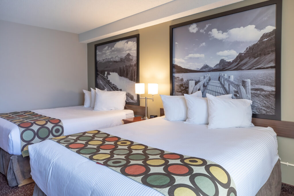 Two double beds, with fresh white bedding,. They each have 3 pillows, and a polka dot throw blanket across the foot of the bed. There are two large black and white photograph print on the wall behind, featuring mountains and lakes, and puffy clouds in the skies. 