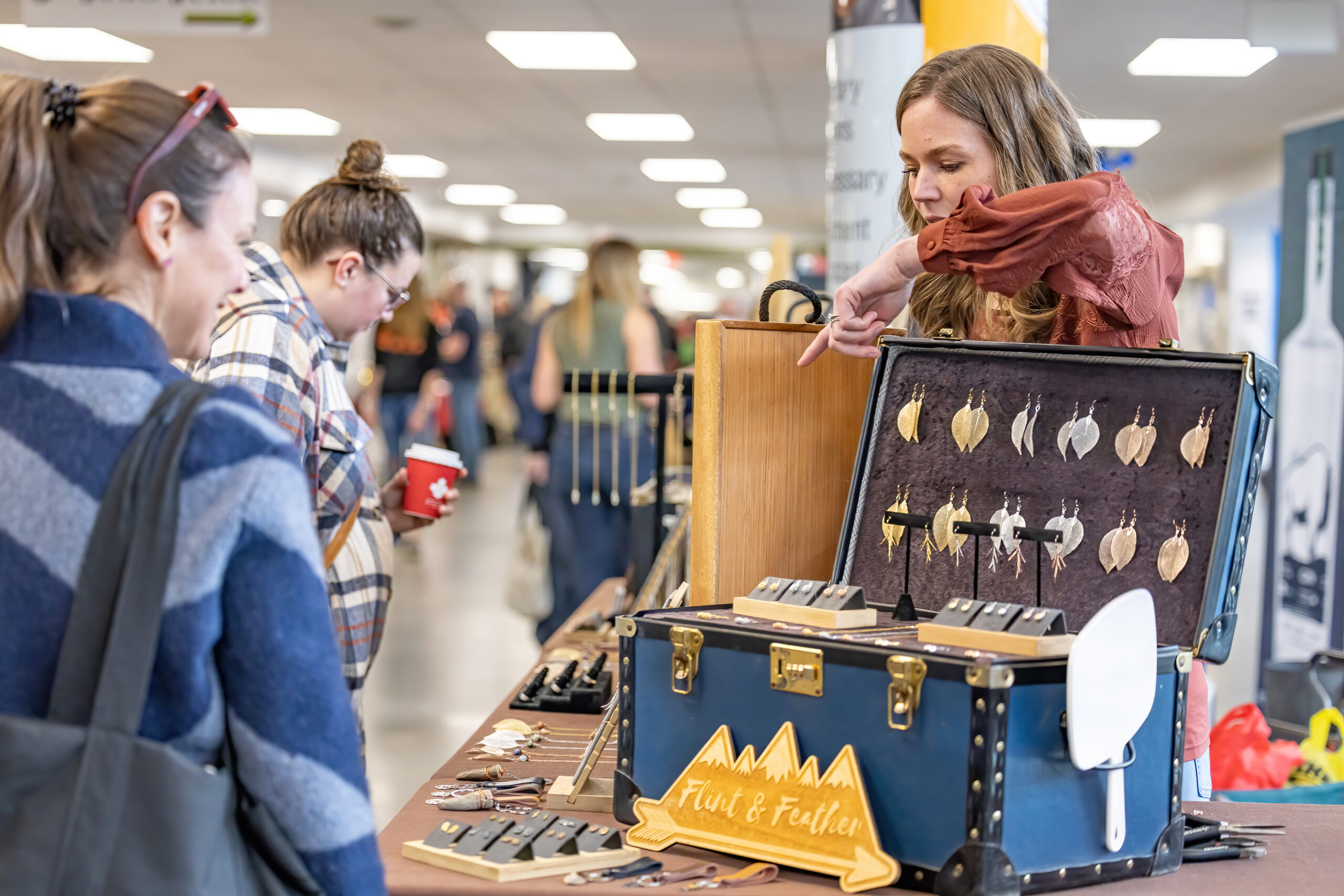 Women look at a jewelry display. Gold and silver leaf earring hang in the lid of a trunk, and the artisan points out more laying on the table.