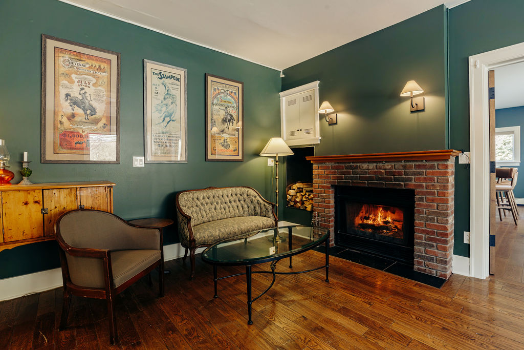 A luxurious room with a brick fireplace. The floors are a rich, medium tone hardwood. The walls are dark green with framed artful vintage posters and sconces. The furniture is vintage, wood and upholstery.