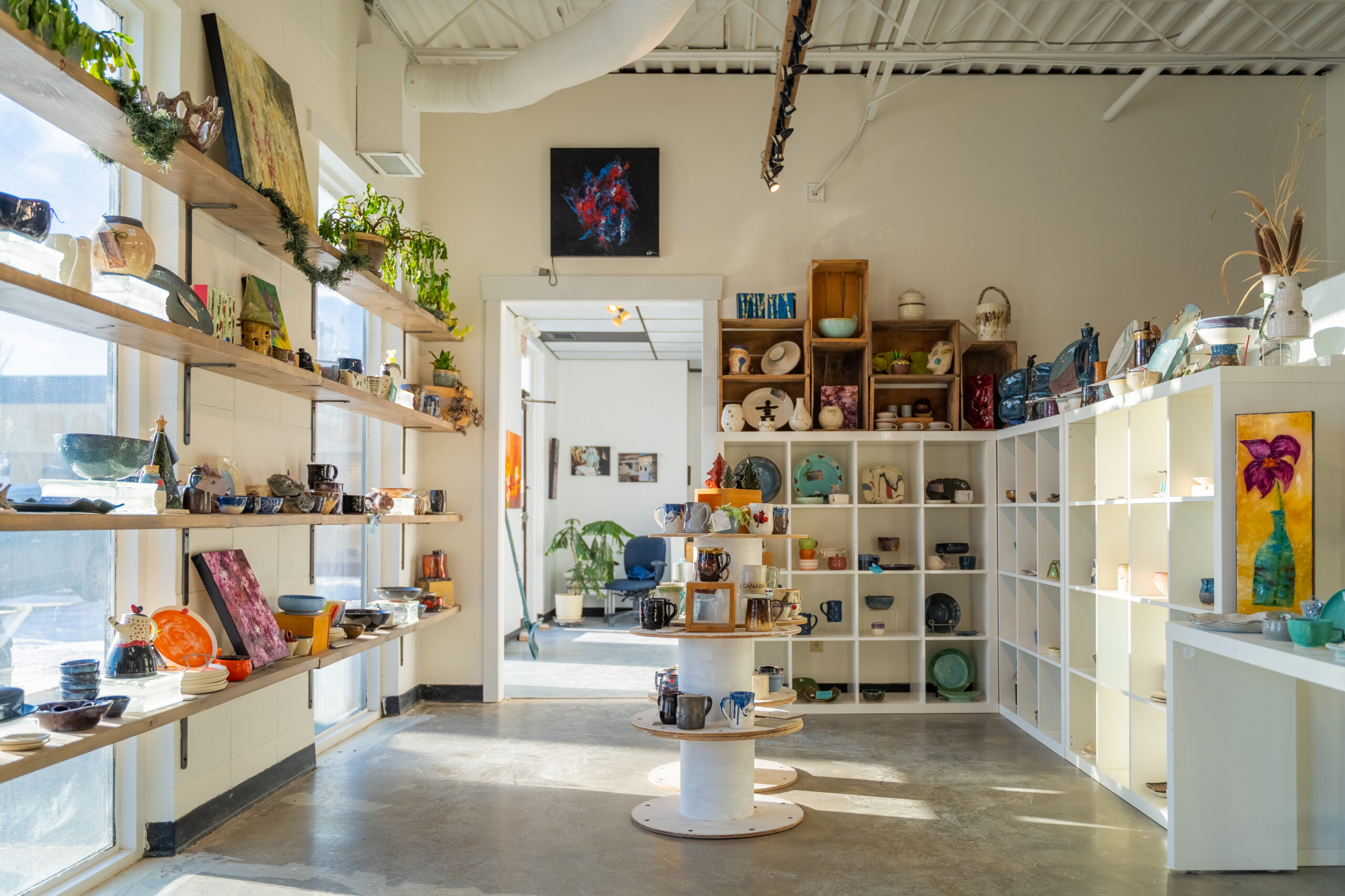 A bright room with sunshine streaming in from the window on the left. The walls are creamy white, the ceiling is high. There are white shelves around the walls and a display table in the middle of the room full of pottery, art, and plants.