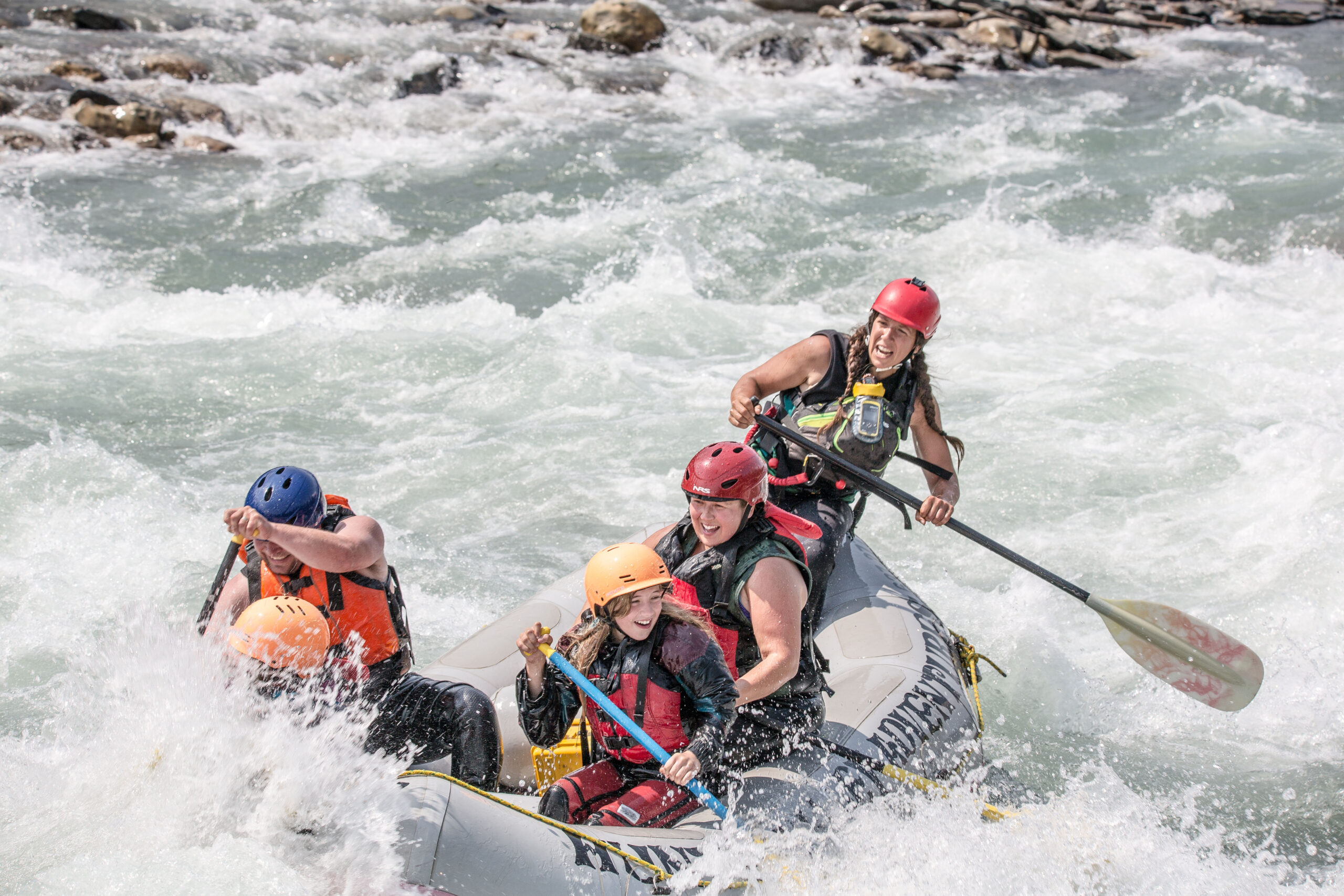 5 people in are paddling through whitewater rapids. They are smiling, some are leaning to balance, and some are getting splashed.