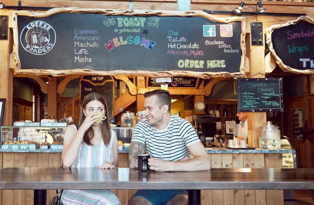 Two people sit at a bar table in a coffee shop. The young woman is sipping her coffee, and the young man is smiling at her. The menu board is behind them. The room is warm and golden, with rustic wood details, colourful writing on the board, and a cozy vibe.