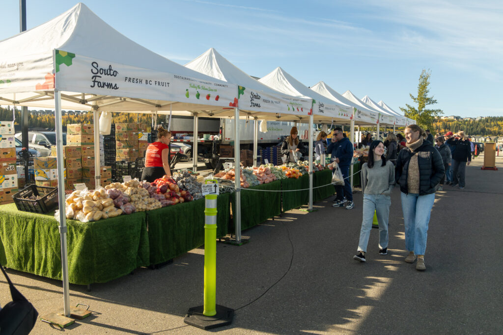 Two young women walk and talk next to a farm stand. Multiple white pop up tents with fresh fruit and veggies from B.C. And more people in the background. It's a sunny day, but the tents offer some shade.