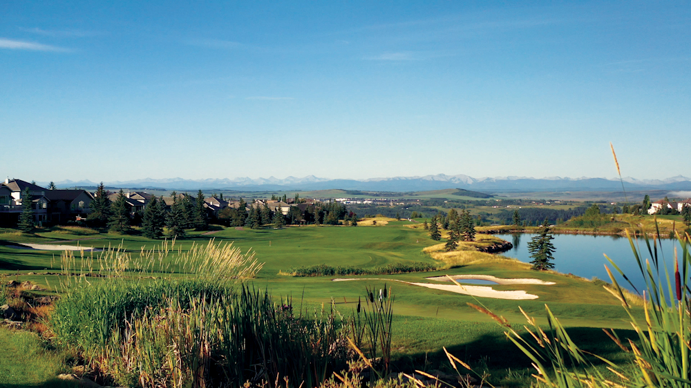 A green golf course under a blue sky. There is a pond on the right, cattails in the foreground, and mountains in the distance.