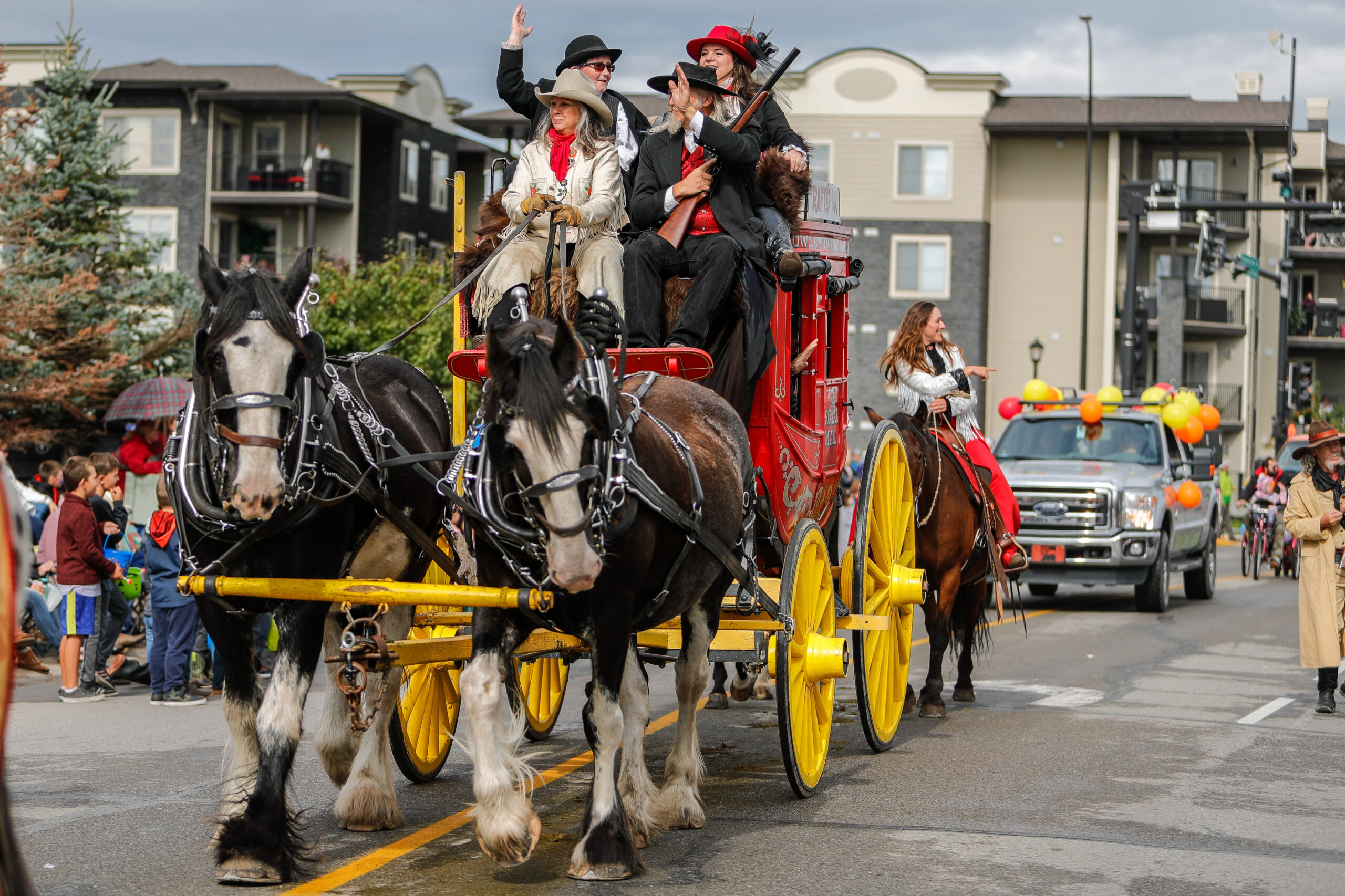 Four people ride on a red carriage with yellow wheels and axels, drawn by two horses. They are part of a parade and waving to a crowd.