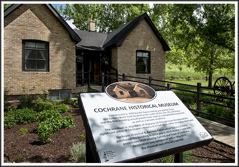 An old brick building sits under a grand tree in dappled sunlight. There is an interpretive sign in front sharing some history of the building. It was built in 1909.