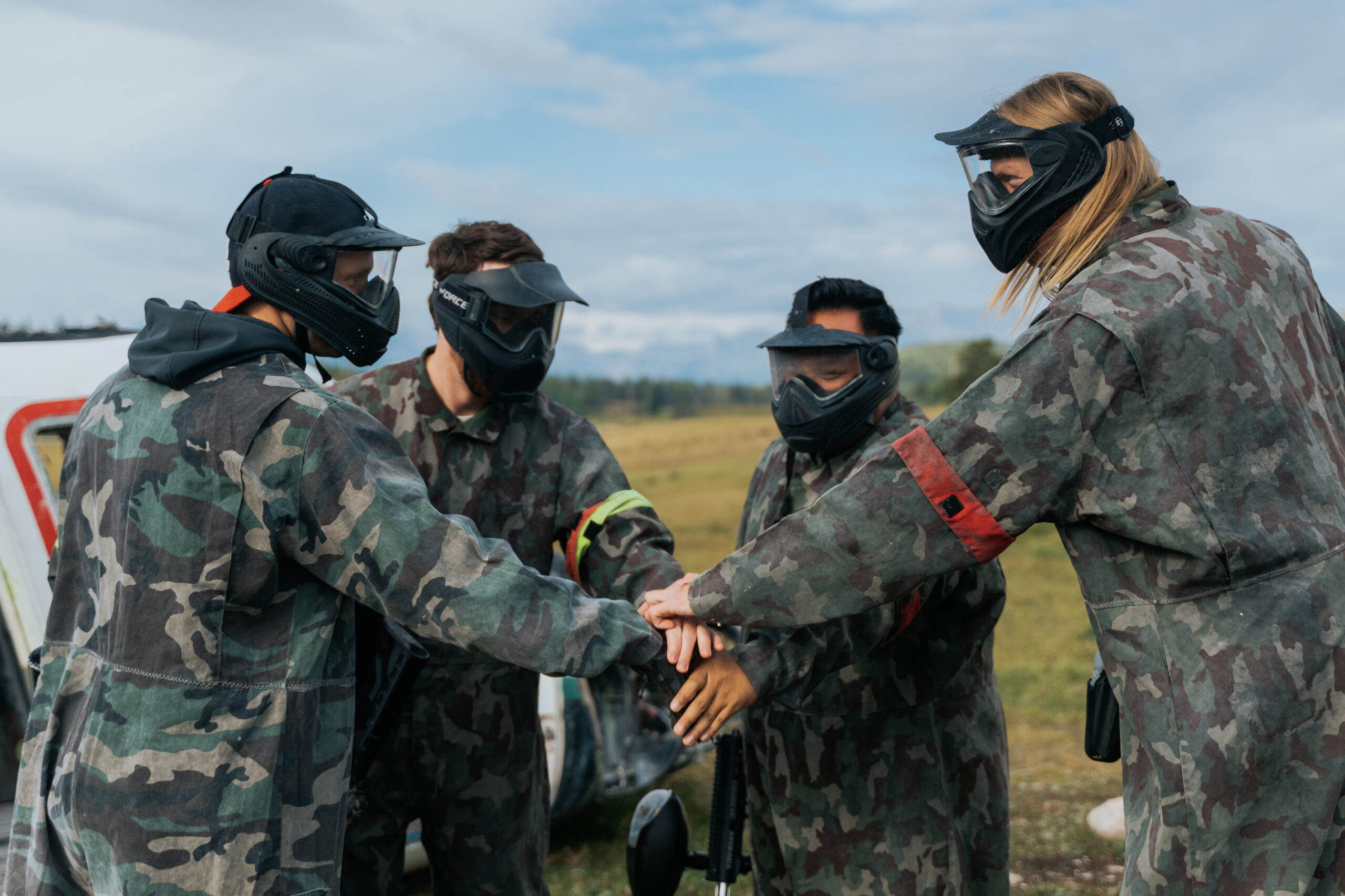 Four men in camo and face shields have their hands in. There is green grass and a blue sky with white fluffy clouds in the background.