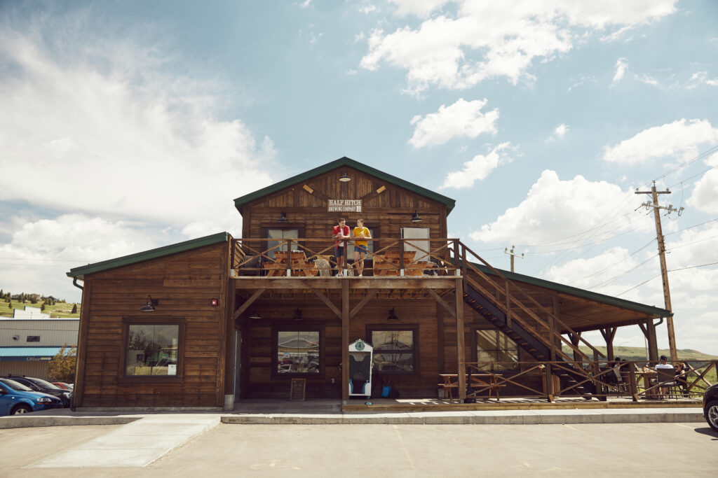 A wooden building against a blue sky. There are people on an upper balcony, and people sitting at tables on a patio on the right. The building has a Western feel.